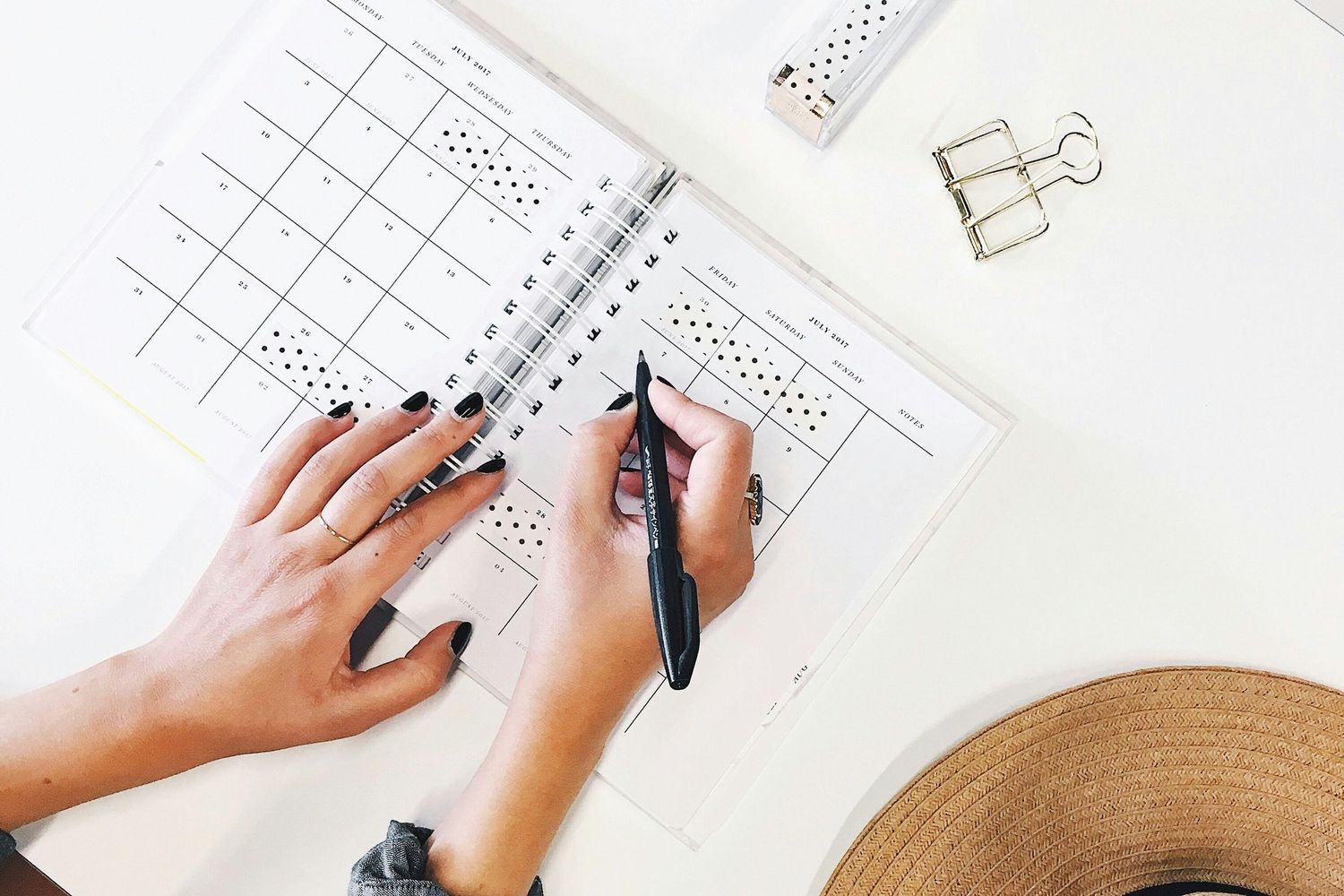 Hands with dark nail polish write in a planner on a white desk with a binder clip and straw hat visible.