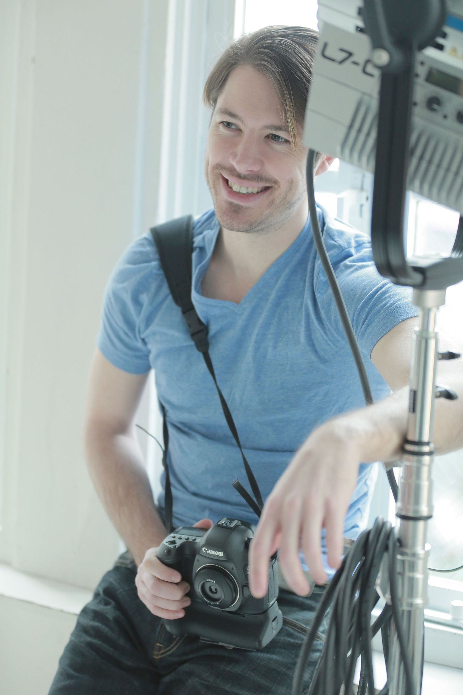 Evan Will, photographer of Sharko Studios wearing a blue t-shirt holding a camera while smiling in a brightly lit studio.