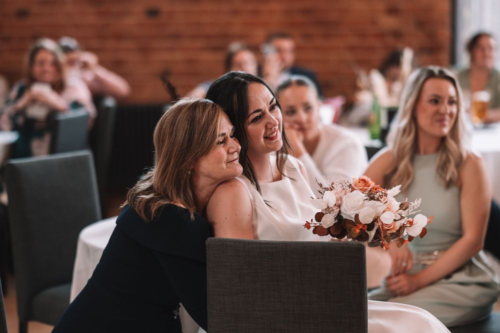 Wedding guests sit in rows of chairs at an indoor ceremony with brick walls and soft lighting.