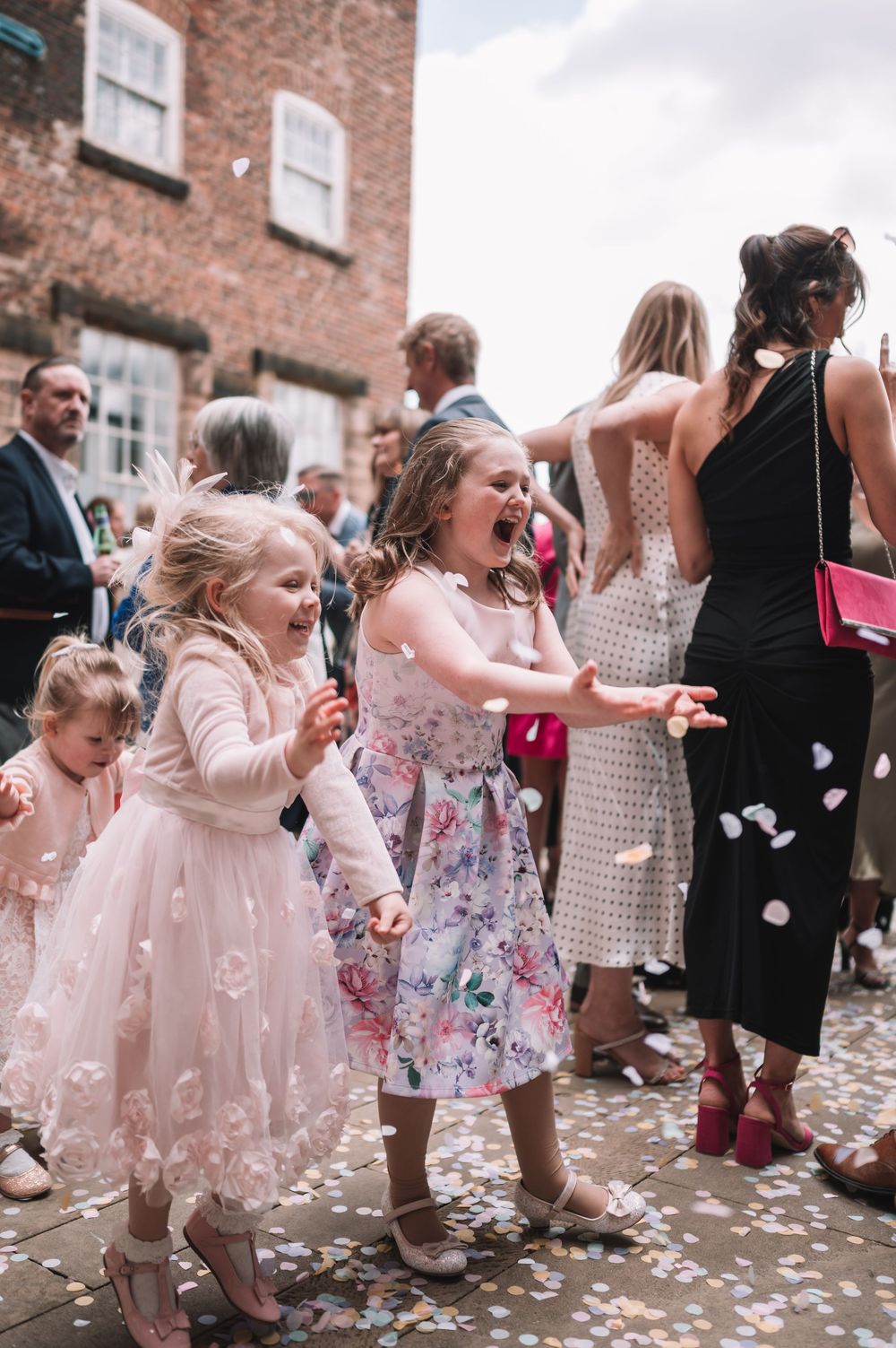 Children in pastel dresses joyfully catch confetti at a celebration outside a brick building.