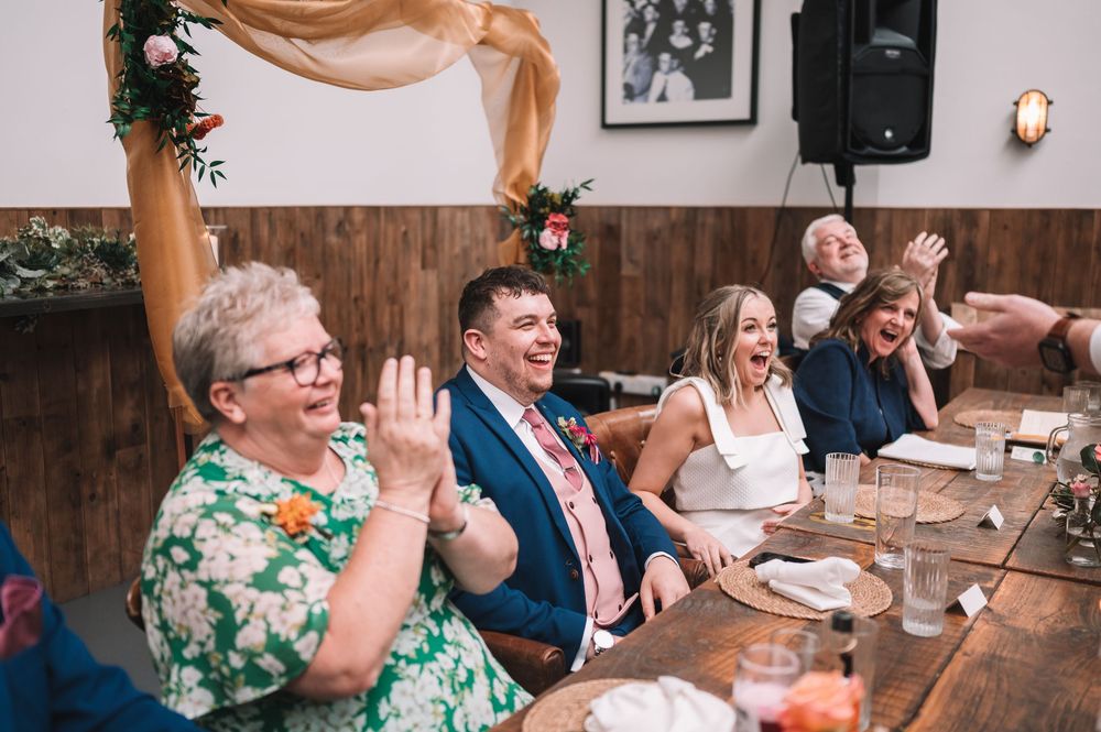 Wedding guests sitting at a long table laugh and wave during reception celebration at rustic venue with wooden walls.