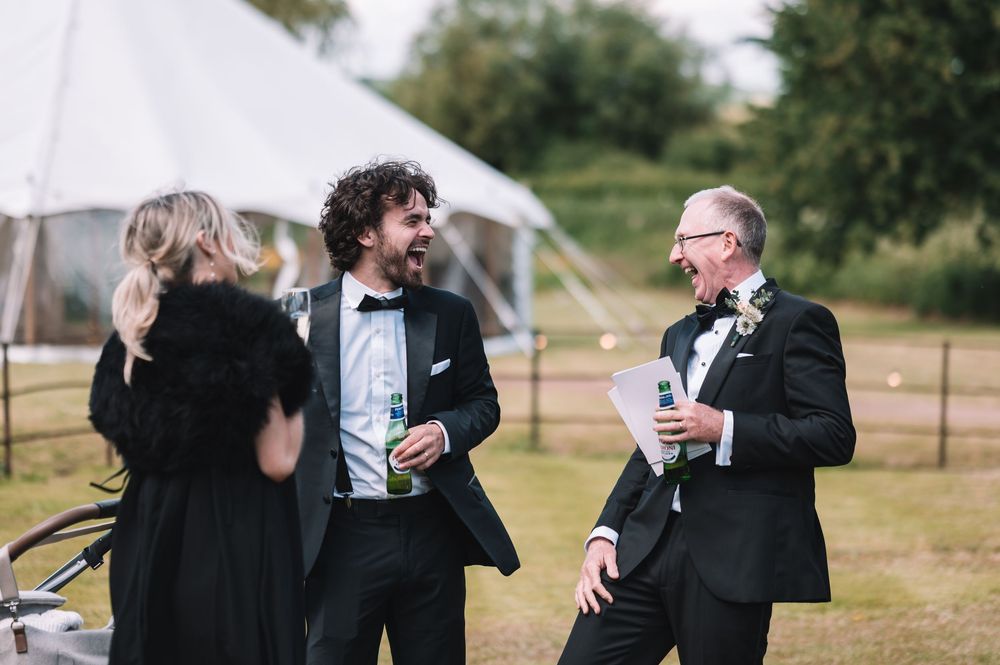 Wedding guests in formal black attire share drinks and conversation outdoors with white tent in background.