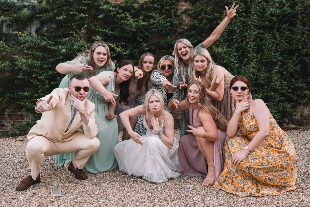 A joyful group in pastel and floral dresses poses together outdoors making silly faces on a gravel path.