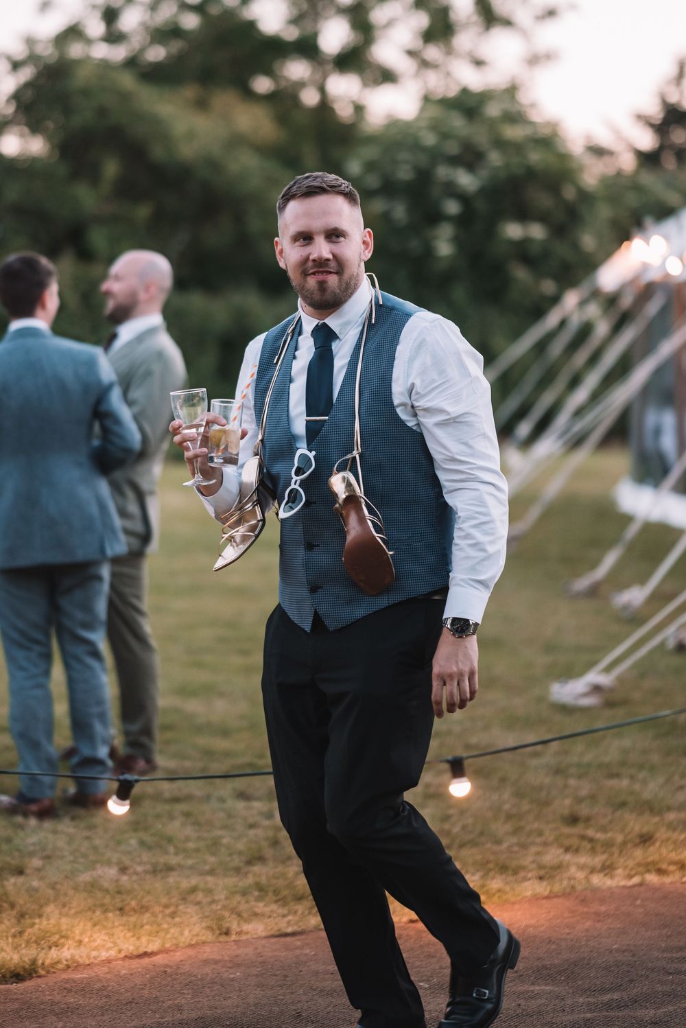 Wedding guest in vest and tie carries champagne glasses at outdoor evening reception with string lights.
