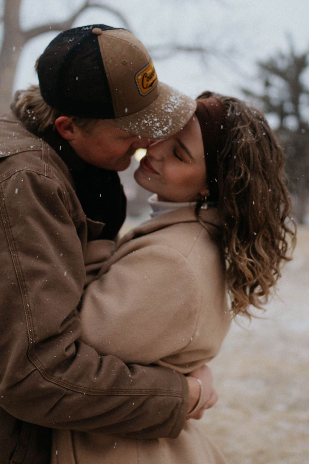 A romantic winter embrace between a couple in warm coats as snowflakes fall around them in Colorado Springs downtown engagement photoshoot