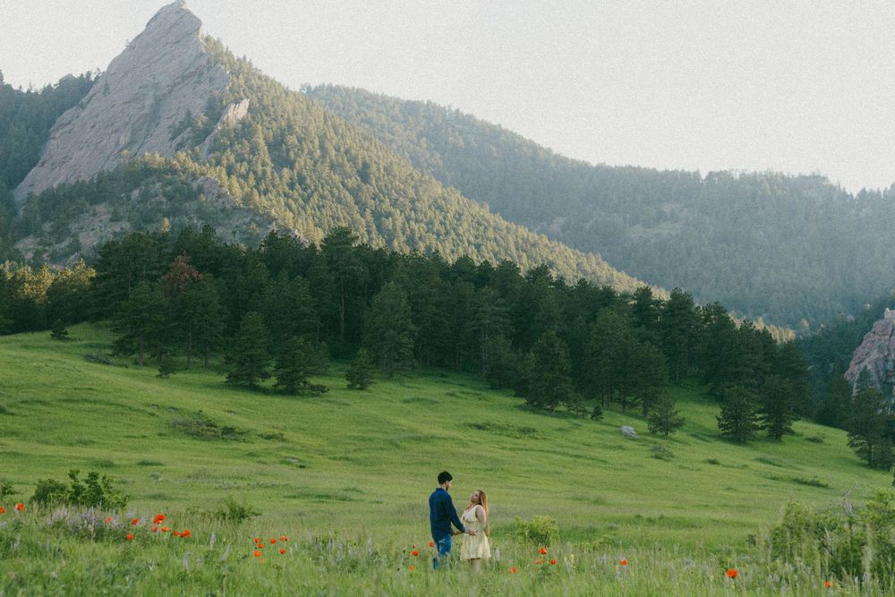 couple walk across a green meadow with dramatic mountain peaks and bloom flowers. Bouder Chautauqua engagement photoshoot