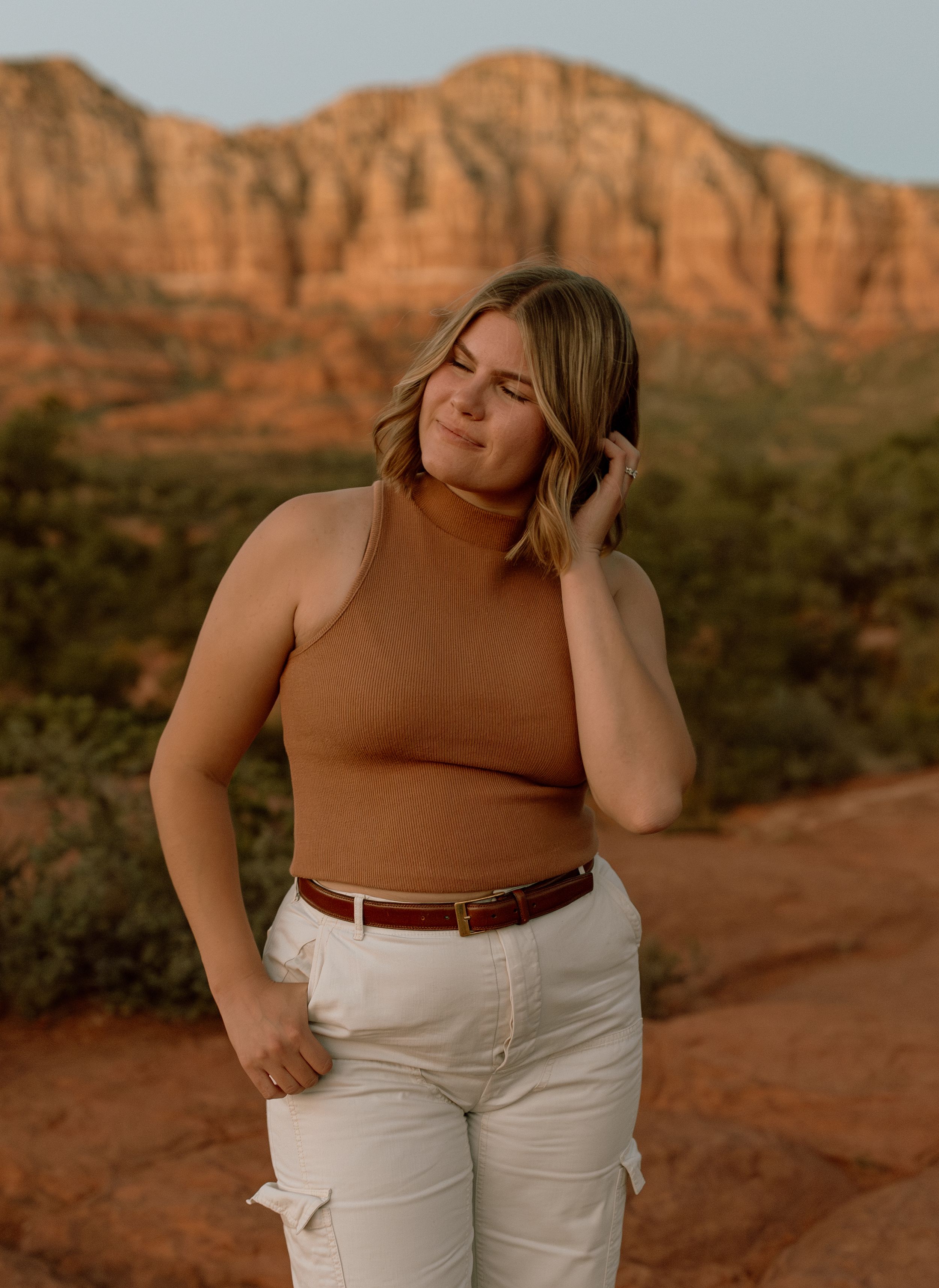 Person wearing brown sleeveless top and white pants poses against red rock formations at sunset.