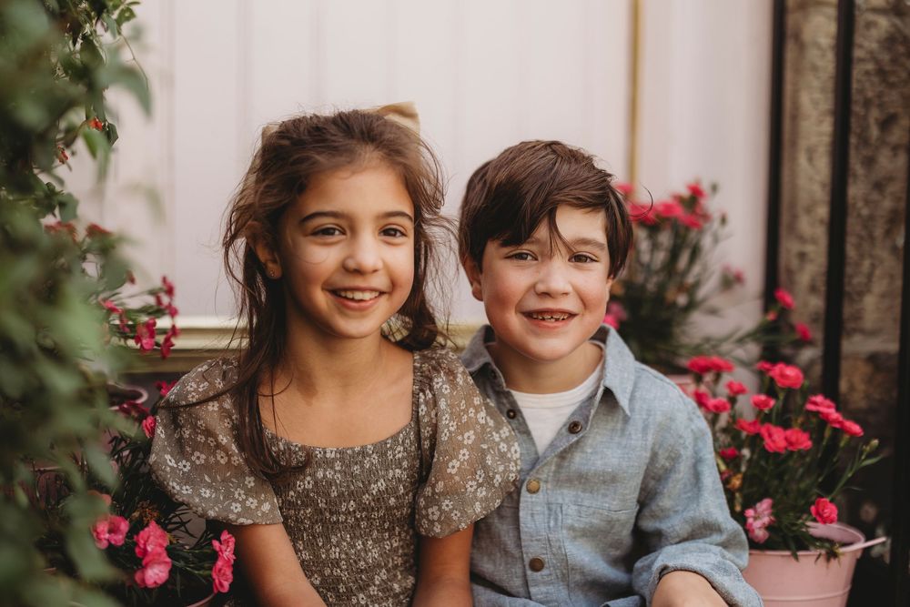 Two children sit and smile together surrounded by pink flowering bushes in an outdoor garden setting.