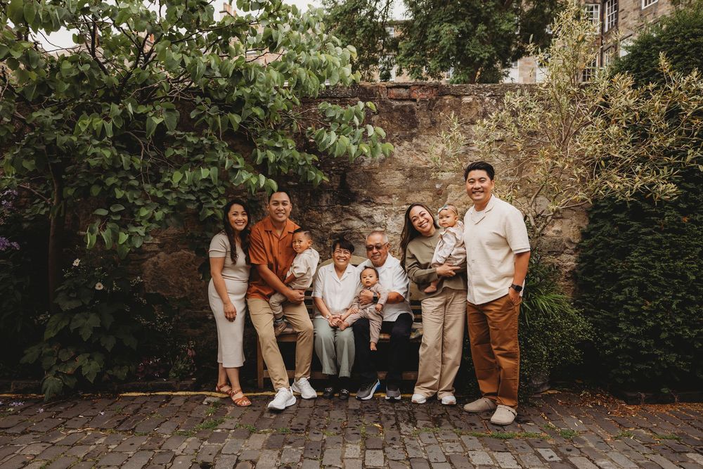 A group in coordinated earth-tone outfits poses together against a stone wall with greenery in an outdoor setting.