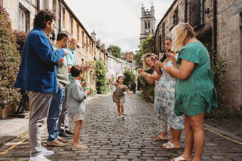 Tourists taking photos along a quaint cobblestone street in Edinburgh with church spire visible in the background.