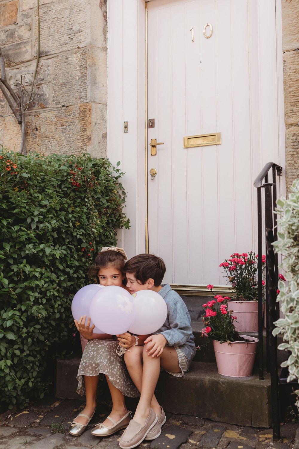 Two children sit on a doorstep holding large white balloons near pink flowers and a garden bush.