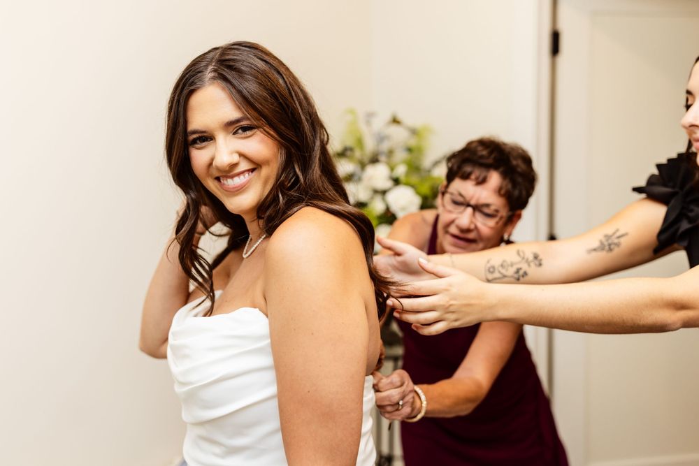 Bride getting ready in white wedding dress while attendant helps with final touches.