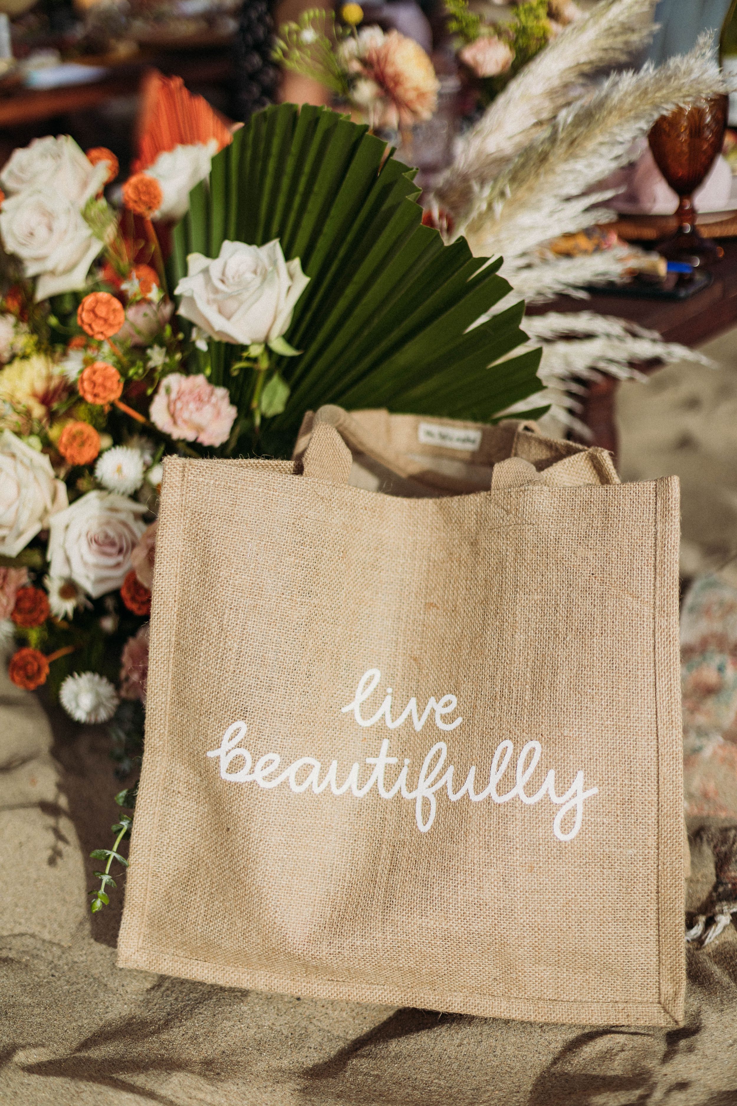 A natural jute tote bag with 'live beautifully' text displayed alongside floral arrangements and palm leaves.