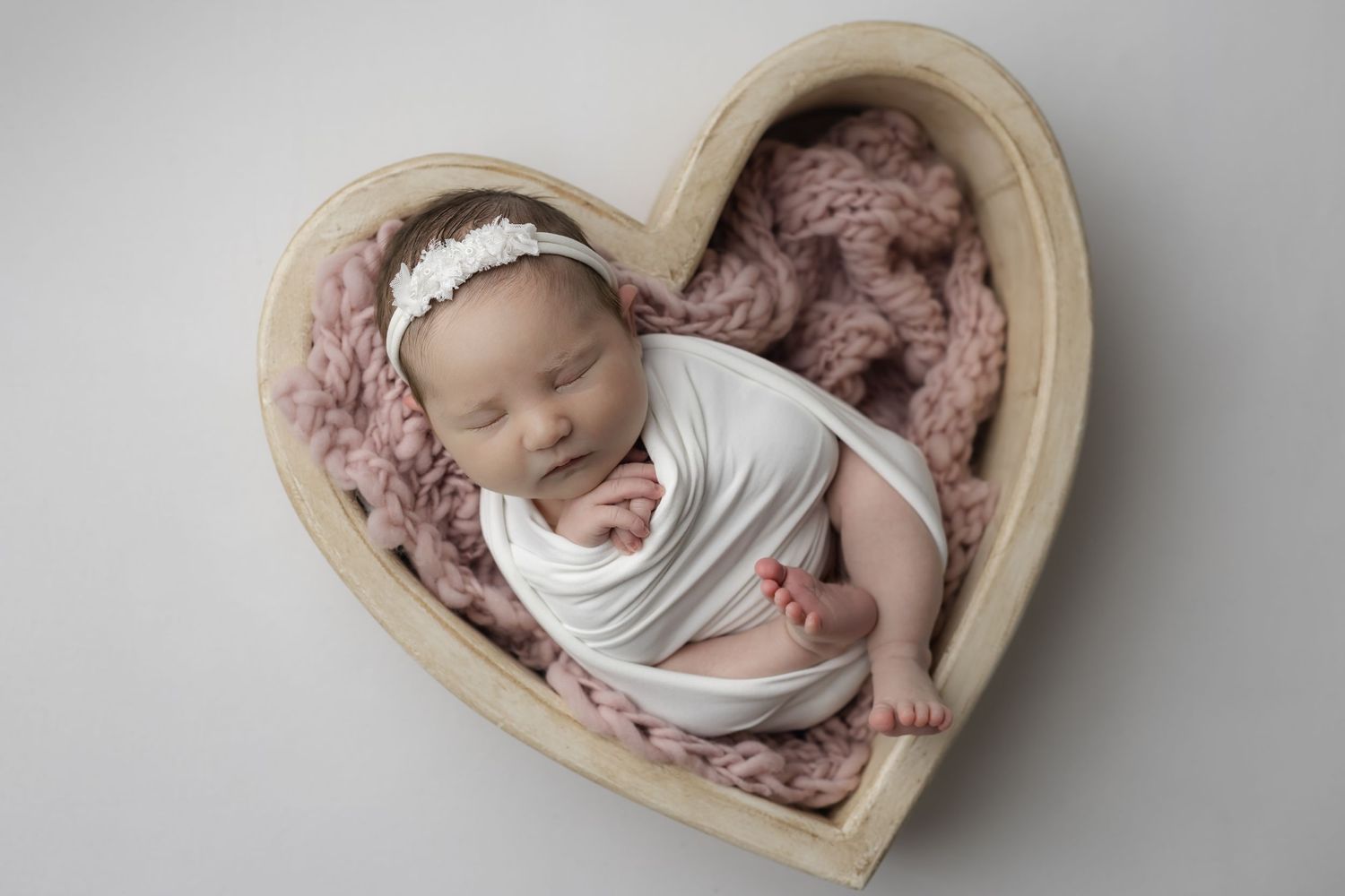 A heart shaped bucket with a swaddled baby girl laying in it wearing white and pink for her studio newborn photoshoot.