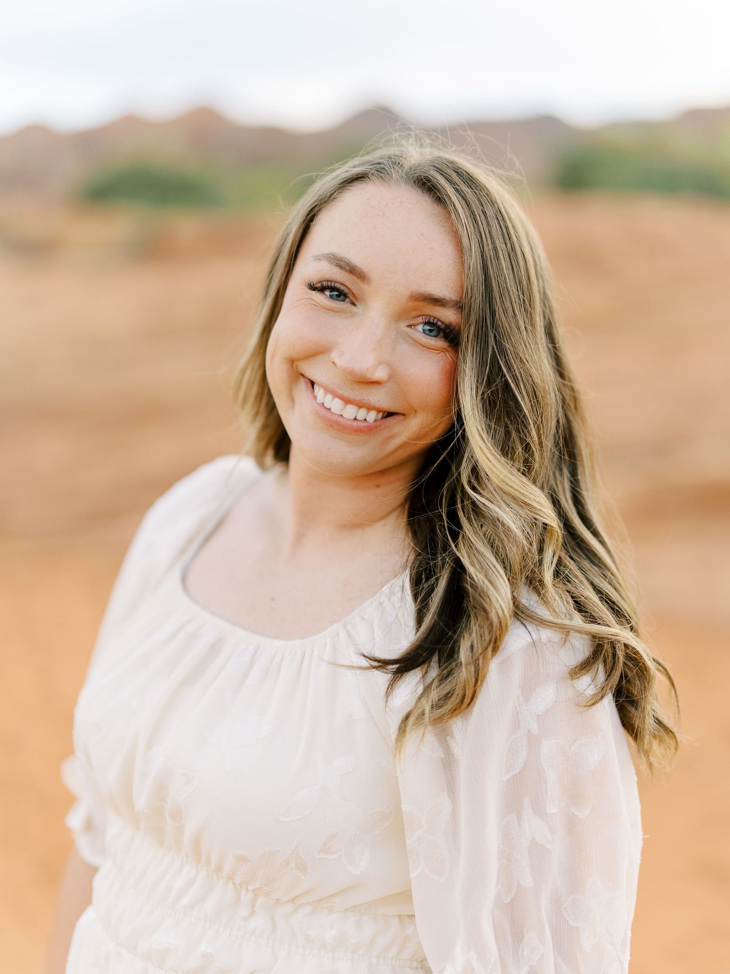 A person in a white dress poses against a backdrop of red desert rocks and rolling hills.