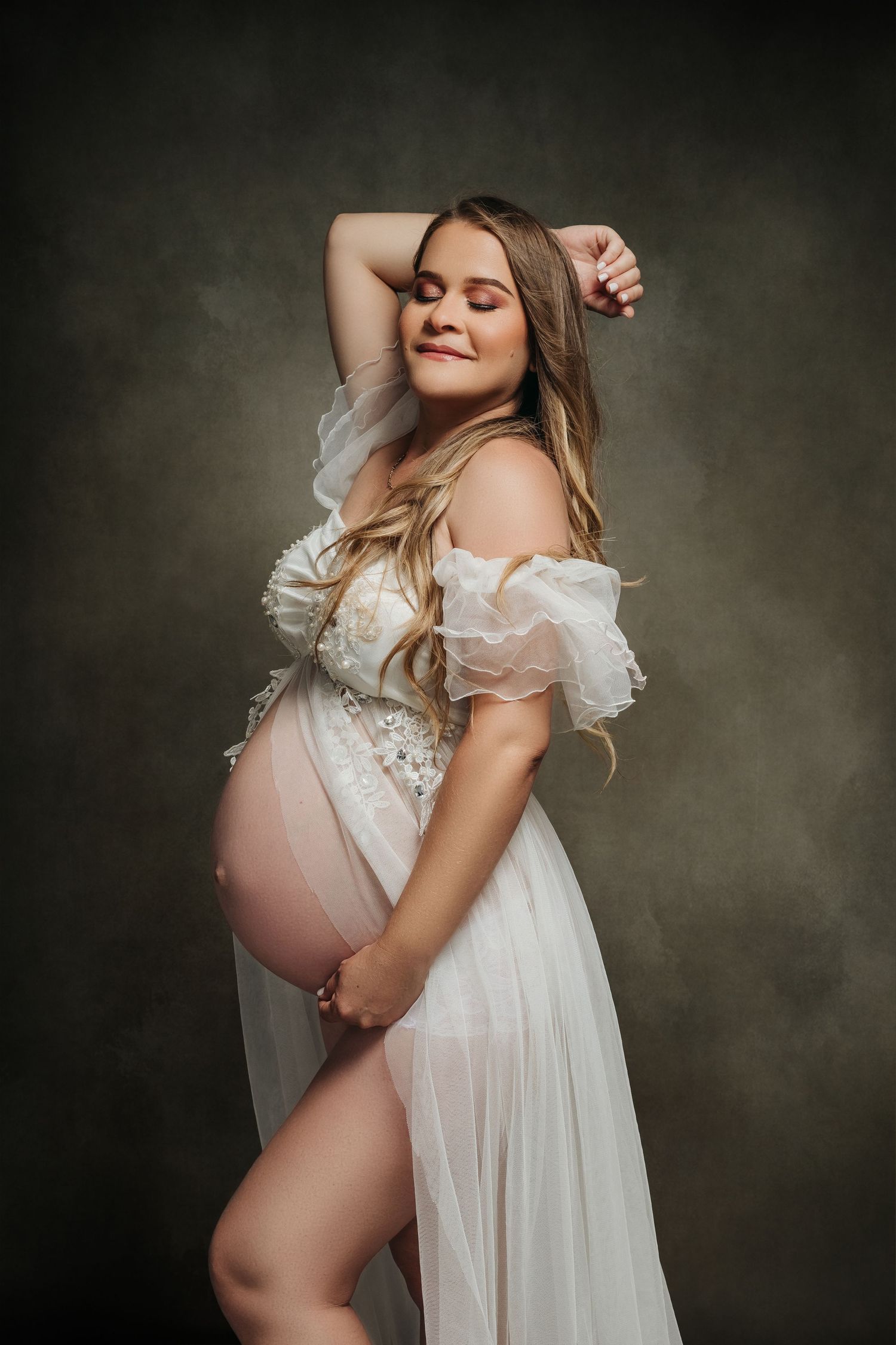 Ethereal maternity portrait with sheer white dress and romantic lighting against dark background.