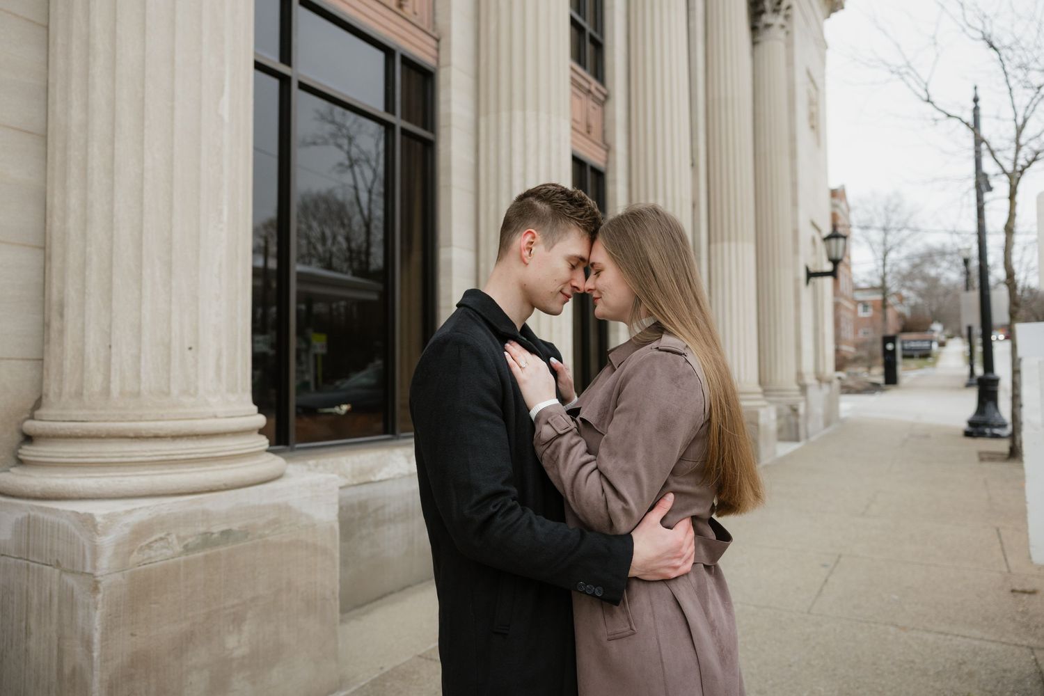 A couple shares an intimate moment outside a building with classical columns, dressed in winter coats.