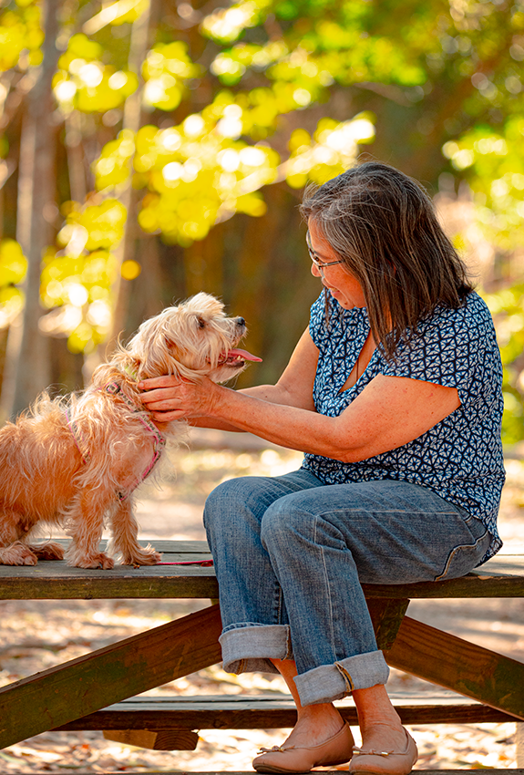 Woman in blue shirt petting small fluffy dog on wooden bench in autumn setting with golden bokeh background.
