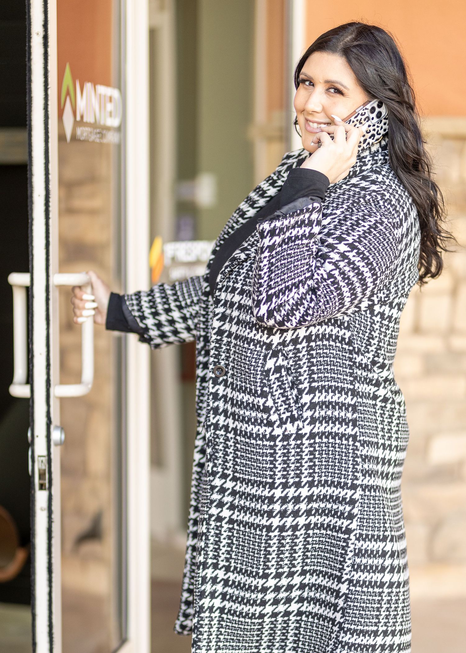 Professional Headshot photo of a woman in a stylish black and white check-patterned coat on the phone outside a business entrance.