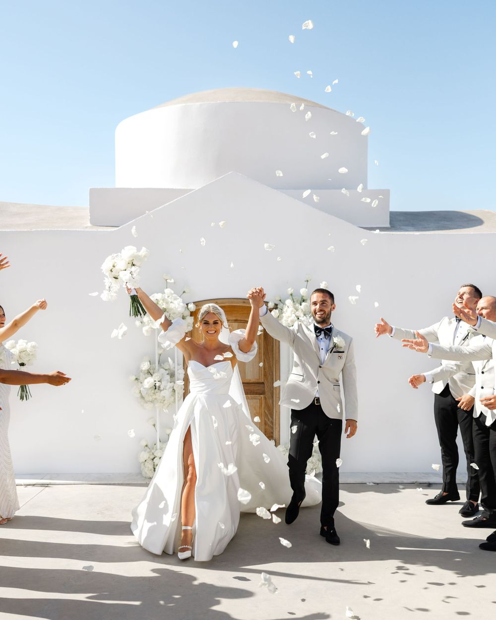 Wedding party throws flower petals in celebration outside a white Mediterranean building.