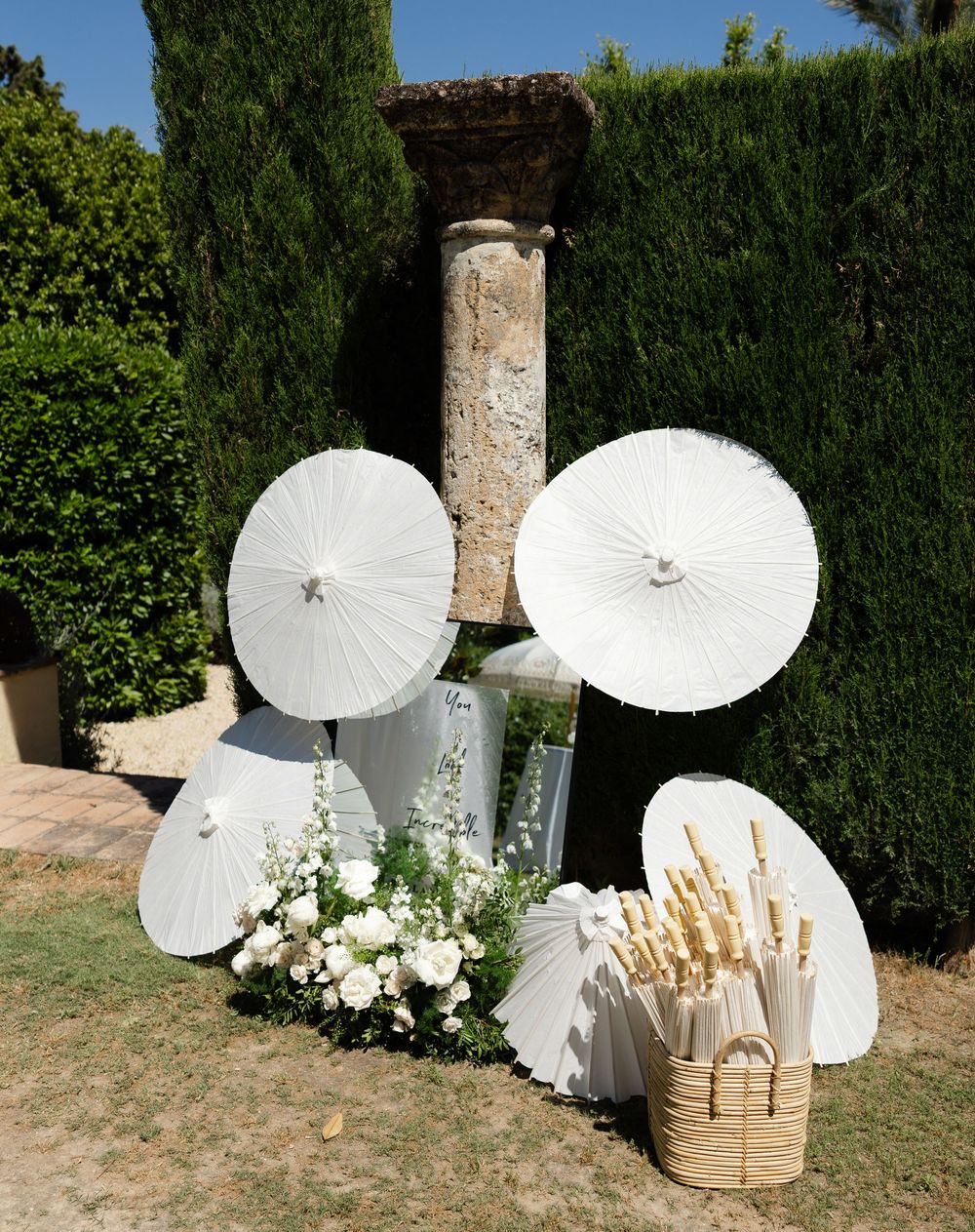 White paper parasols and floral arrangements create an elegant outdoor wedding display against a green hedge.