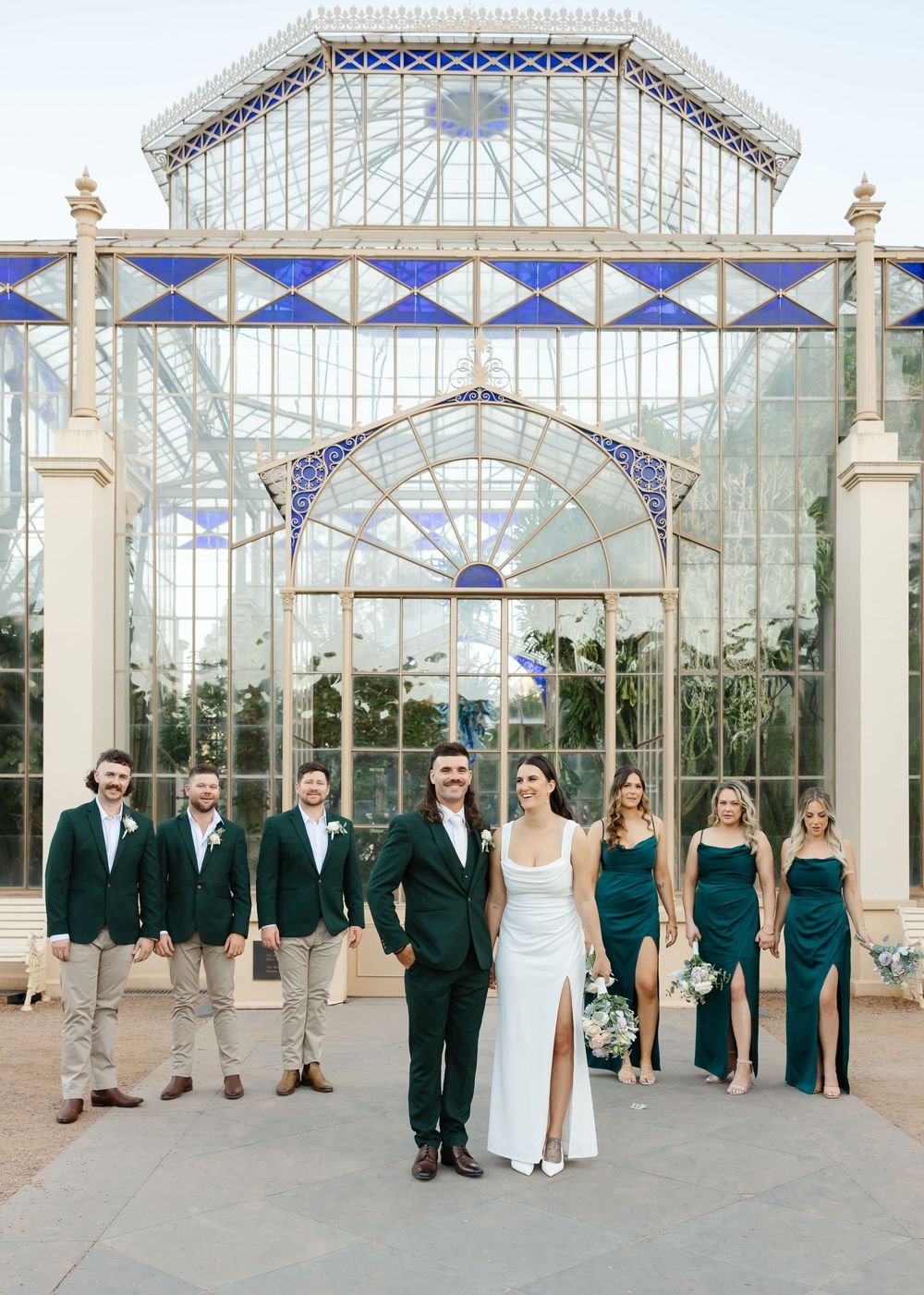Wedding party poses in front of ornate glass conservatory with blue and white geometric architectural details.