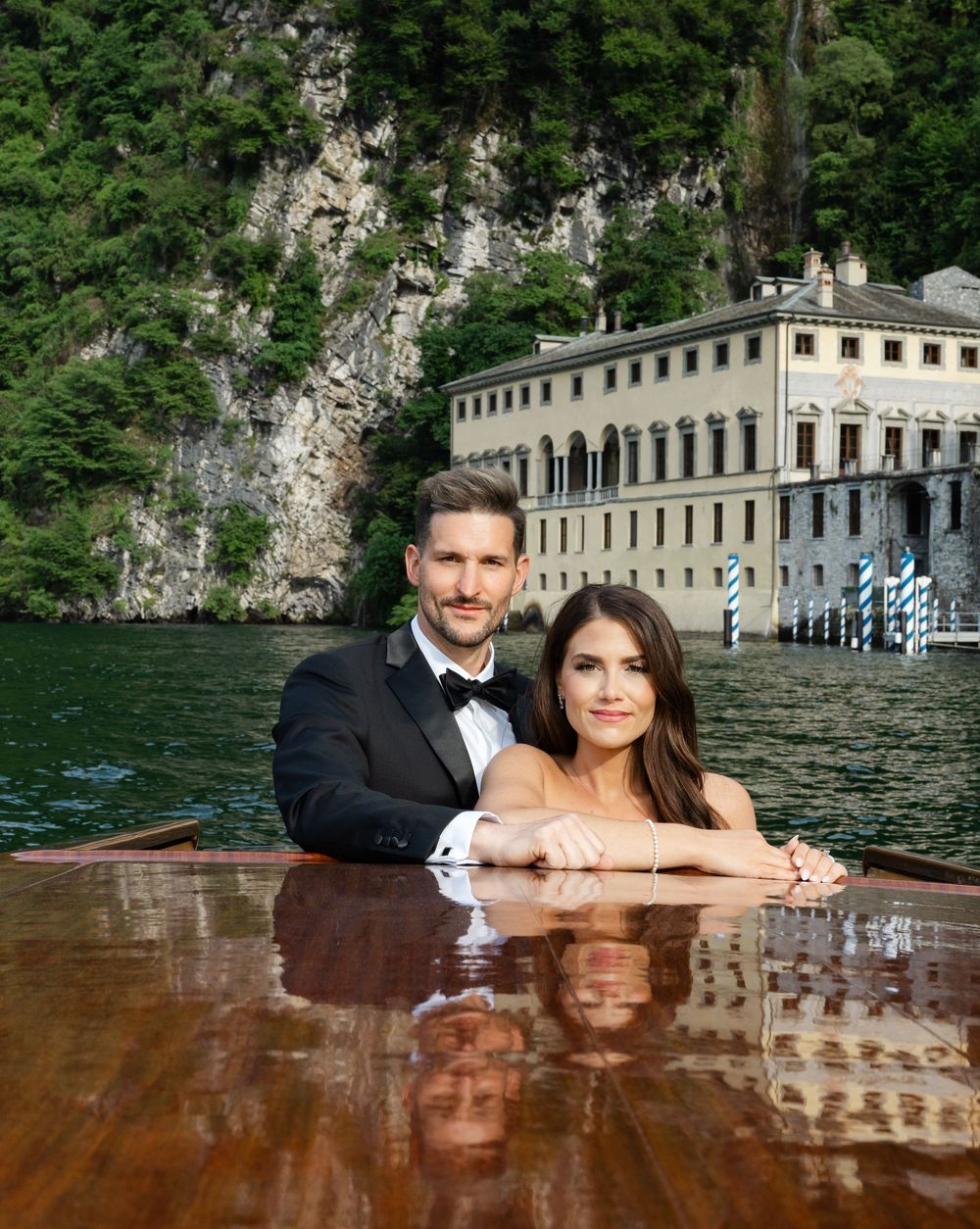 A couple poses together on a polished mahogany table by Lake Como with historic Italian villa in background.
