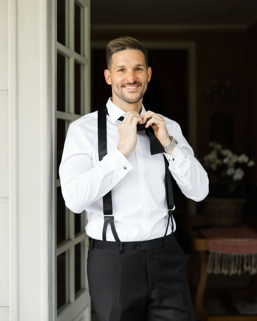A person in a white dress shirt and dark suspenders smiles warmly while standing near white french doors.