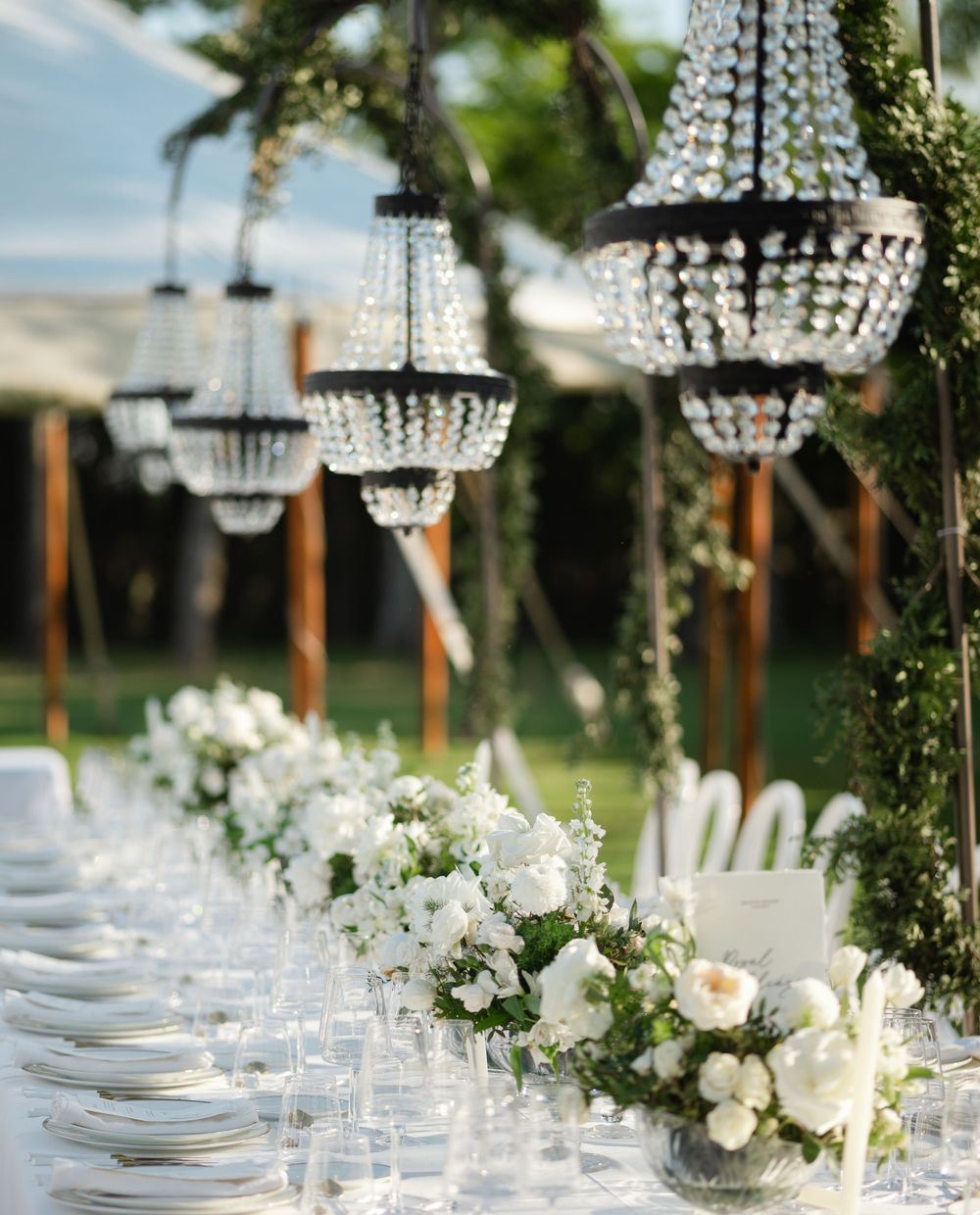 Crystal chandeliers hang over an elegantly set white dining table with floral centerpieces.