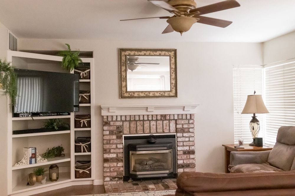Cozy living room featuring brick fireplace, ornate gold mirror, built-in shelving, and ceiling fan.