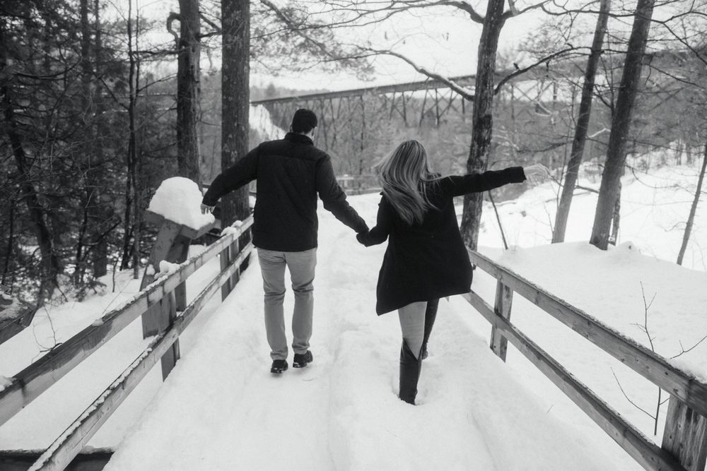Black and white photo of couple walking hand in hand on snowy bridge.