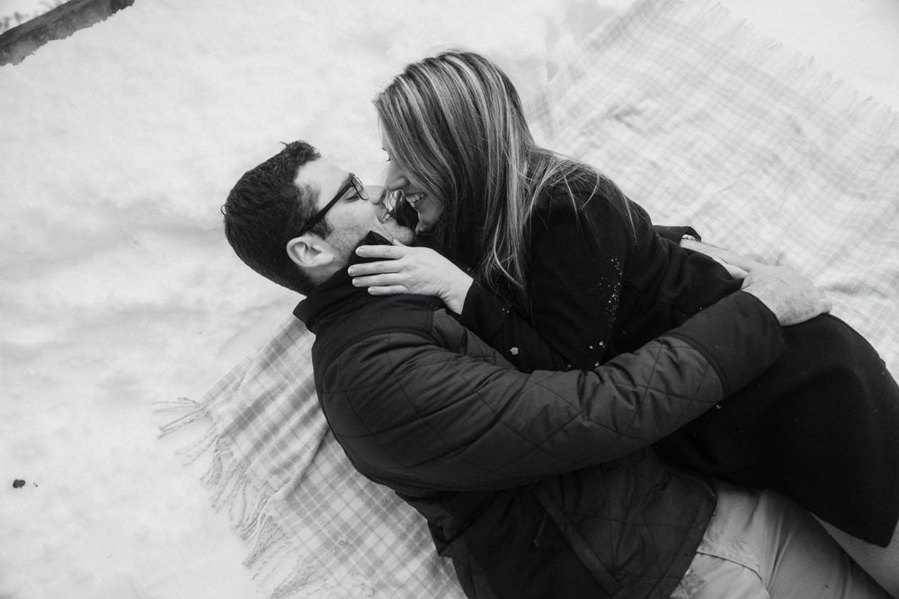Black and white photo of couple lying in snow sharing intimate moment.