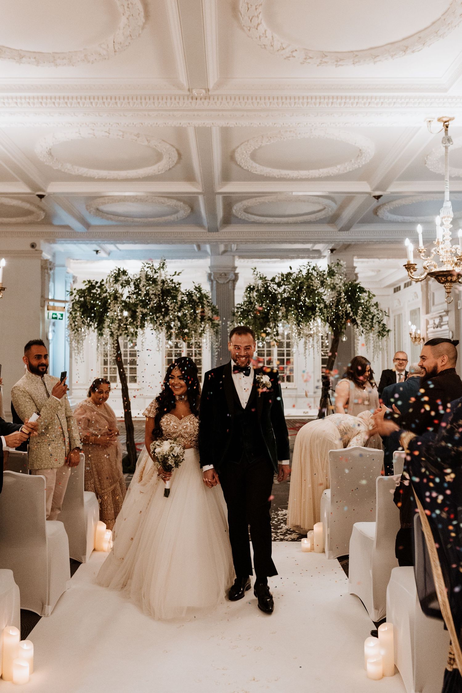 Elegant wedding ceremony in a white room with ornate ceiling, greenery arches and candle-lined aisle at indoor venue.