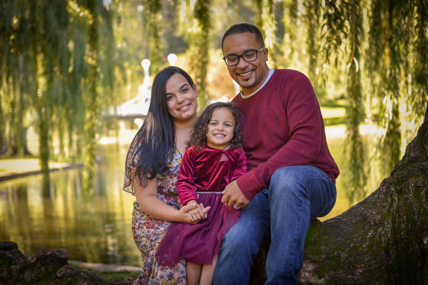 A family of three posing together under a weeping willow tree near a pond.
