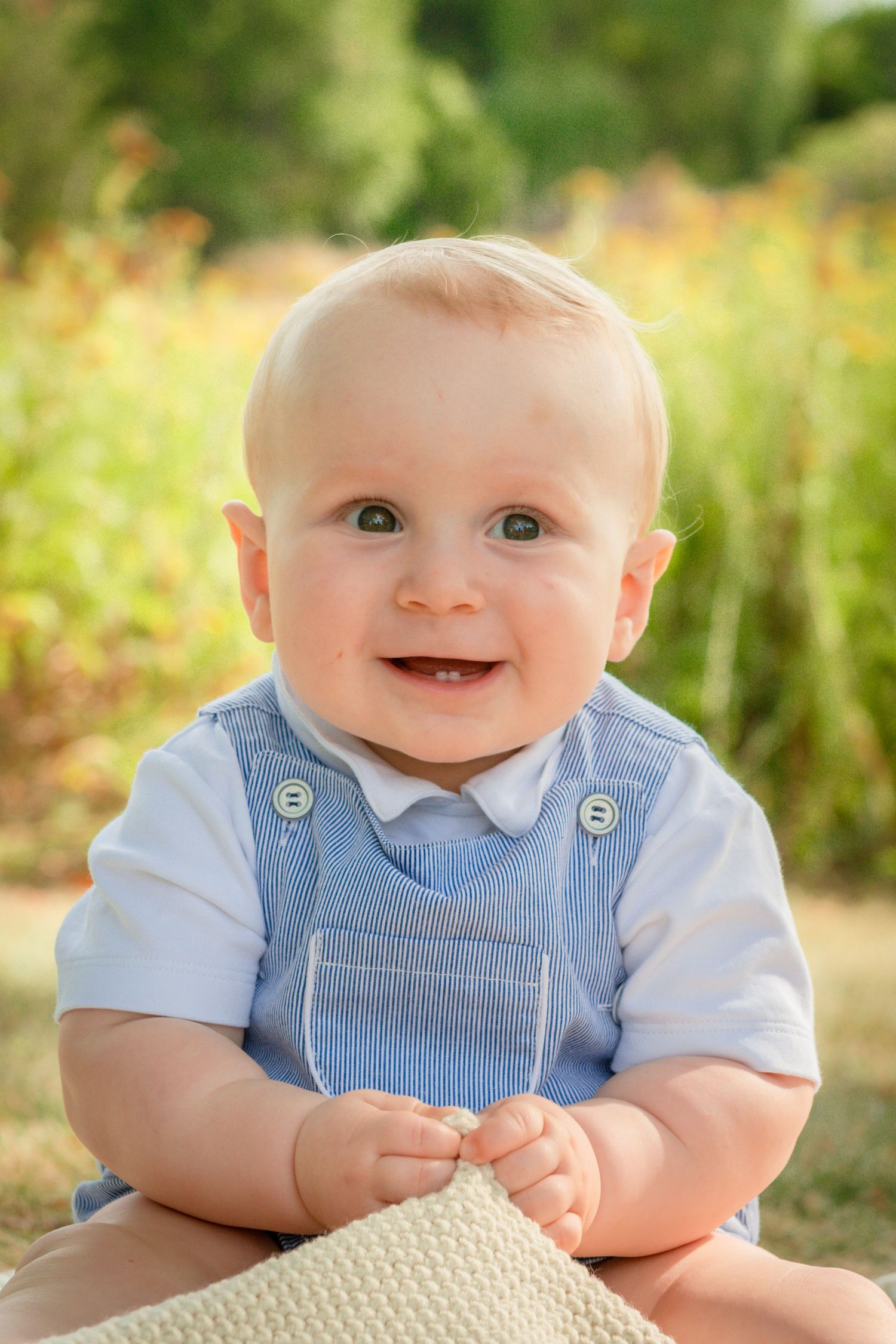 A baby in blue and white striped overalls smiling at the camera outdoors.