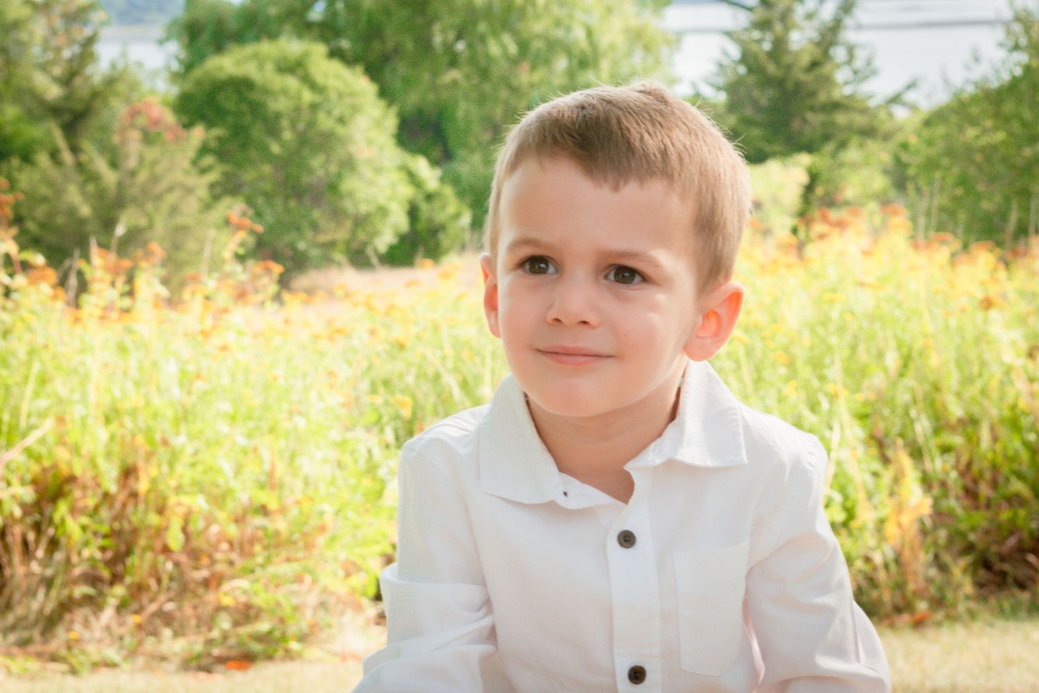A young child in a white polo shirt poses in front of a natural summer backdrop.