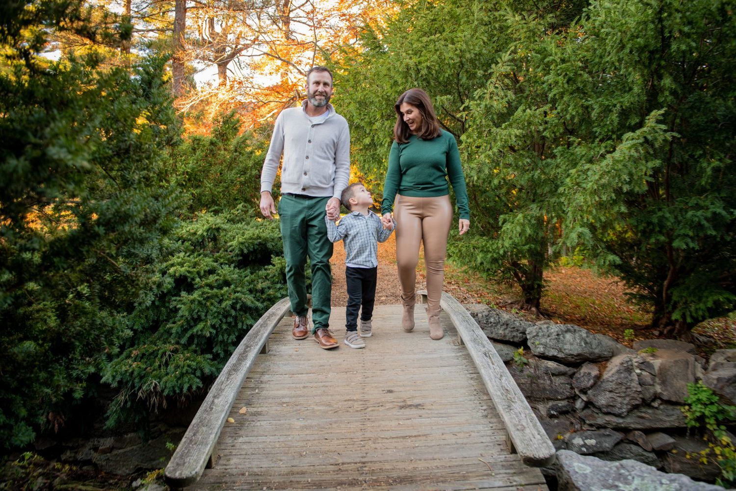 A family walks together across a wooden footbridge surrounded by fall foliage.