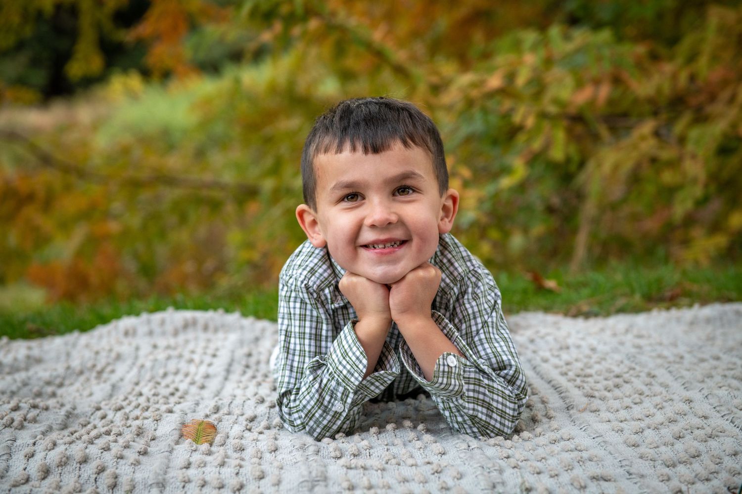 A child lying on a blanket outdoors smiles at the camera during an autumn photo session.