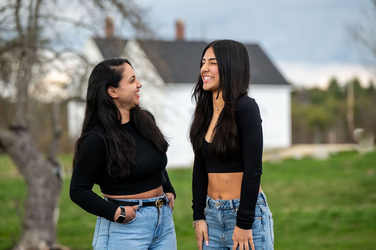 Two sisters in cropped tops and jeans share a candid moment in front of a white farmhouse. New Hampshire Family Photography Serving