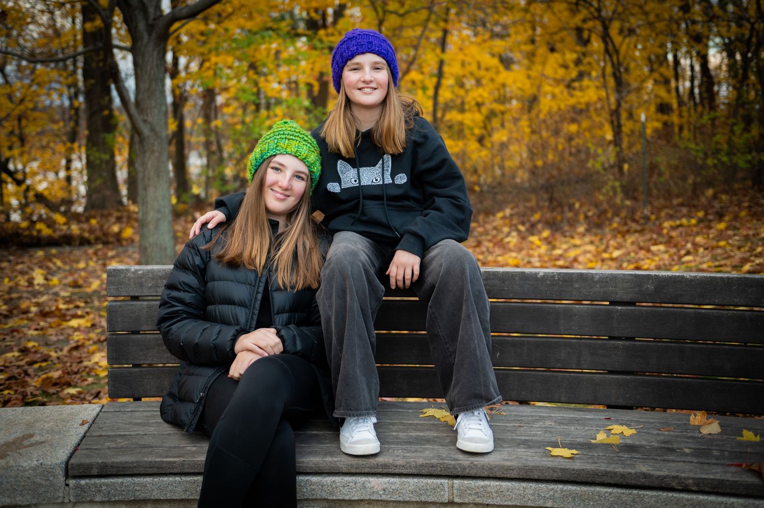 Two sisters in winter gear sitting on a wooden bench surrounded by golden fall leaves.