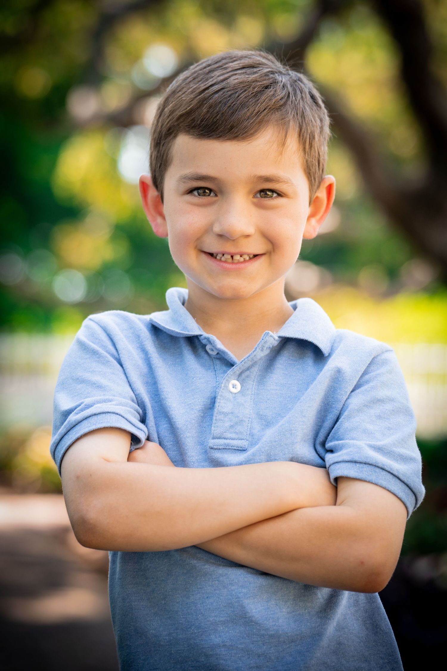 A child in a light blue polo shirt poses with arms crossed against a blurred garden background. New Hampshire Family Photography