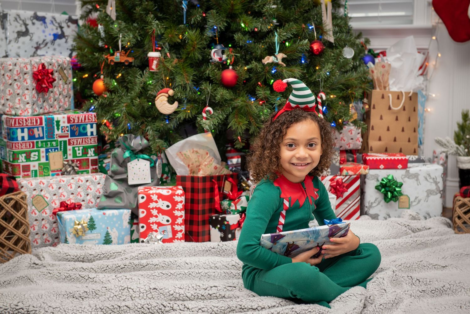 A child in green pajamas sitting by a Christmas tree surrounded by wrapped presents.