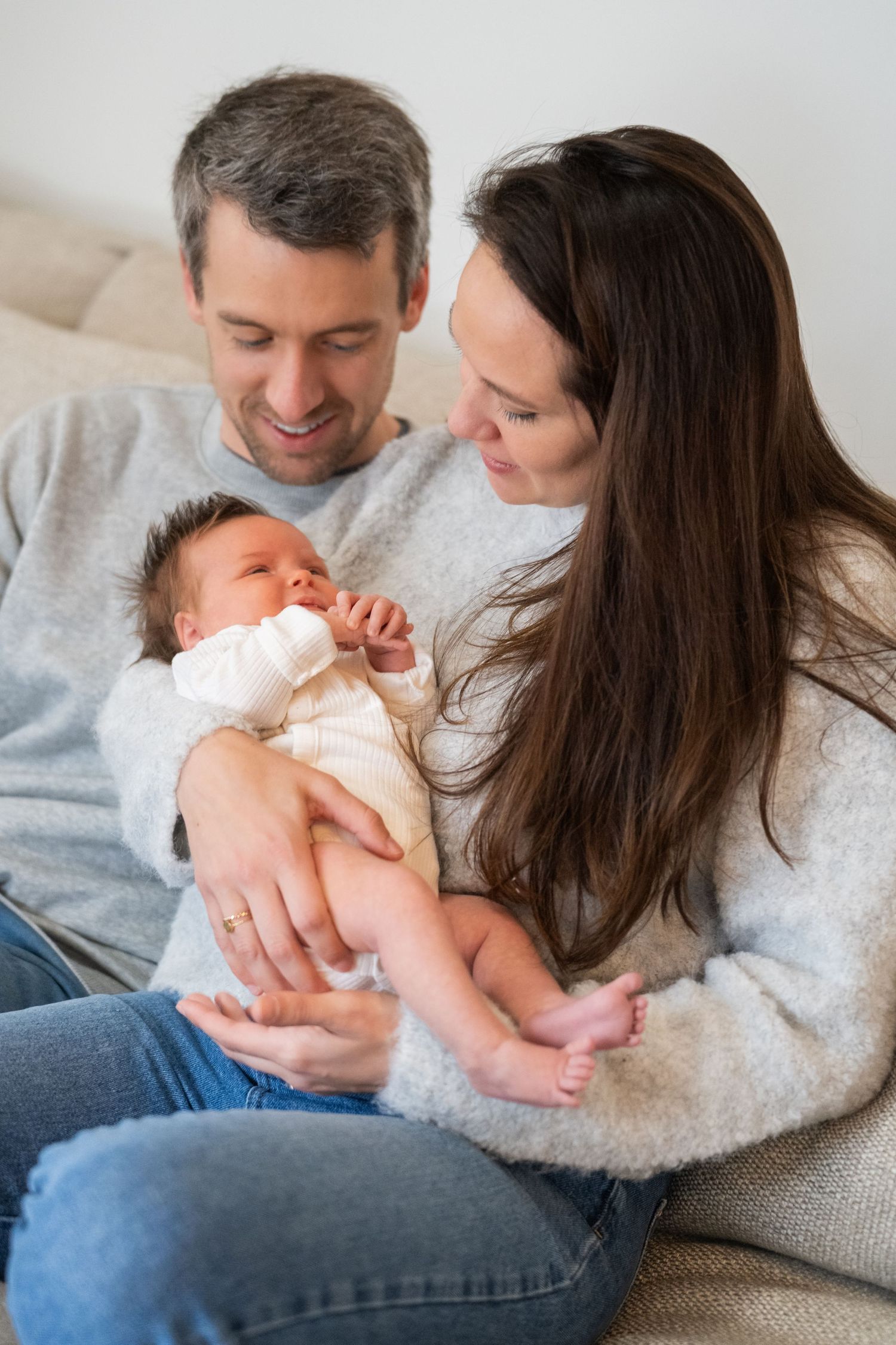 Parents lovingly gaze at their newborn baby wearing white while sitting together on a couch.