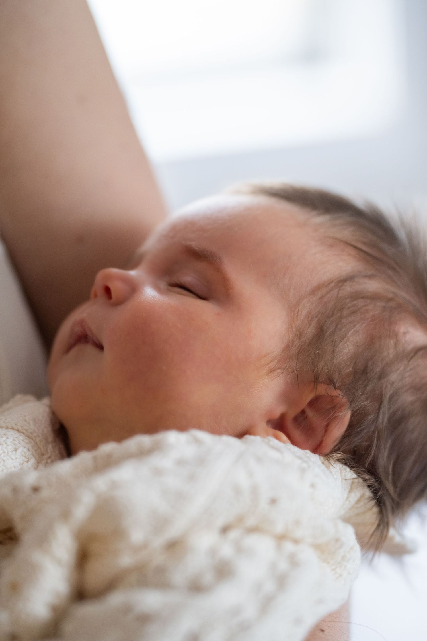 A sleeping baby rests peacefully against white textured bedding in soft natural lighting.