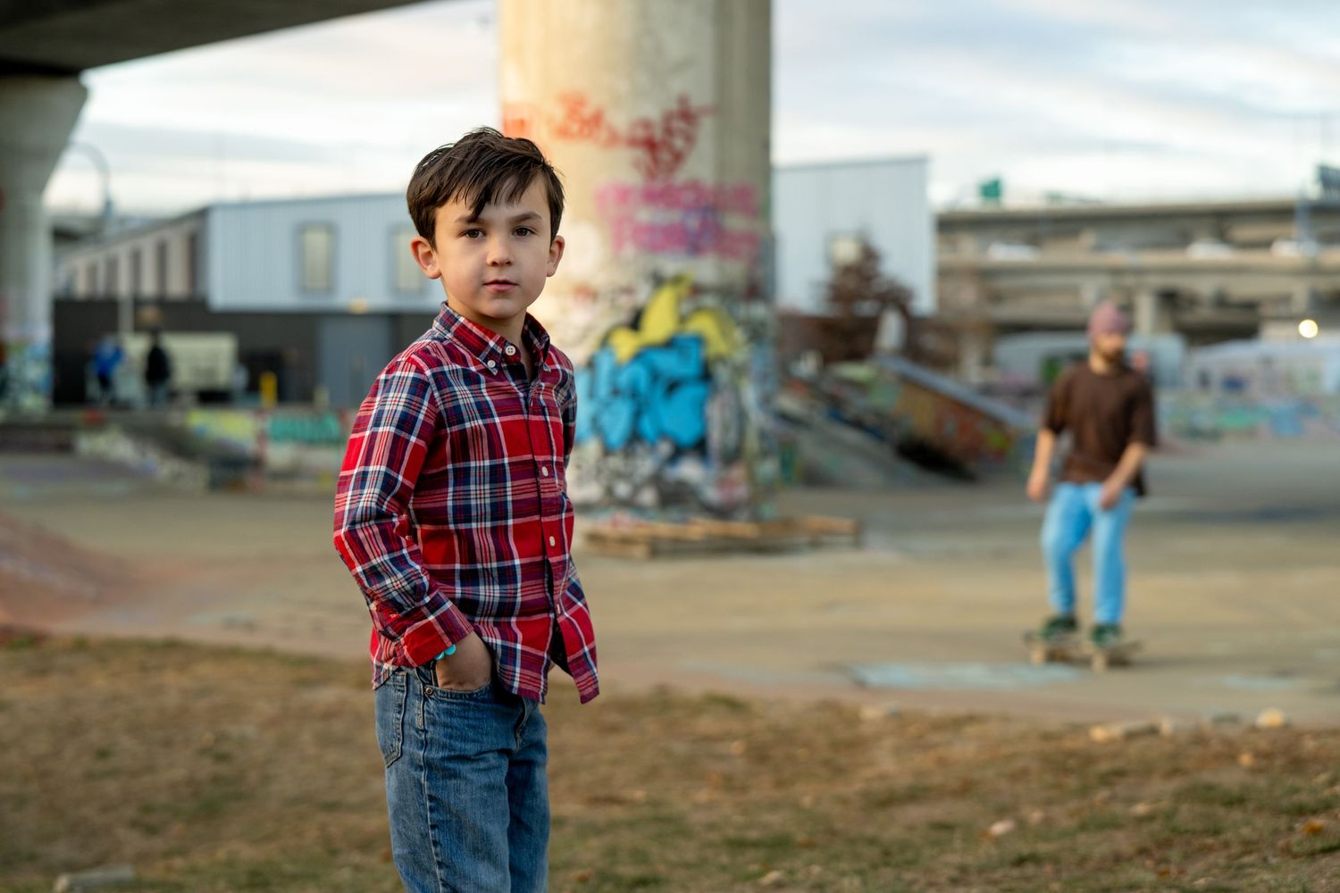 A young child in a red plaid shirt and jeans stands in an urban setting with graffiti and buildings in the background.
