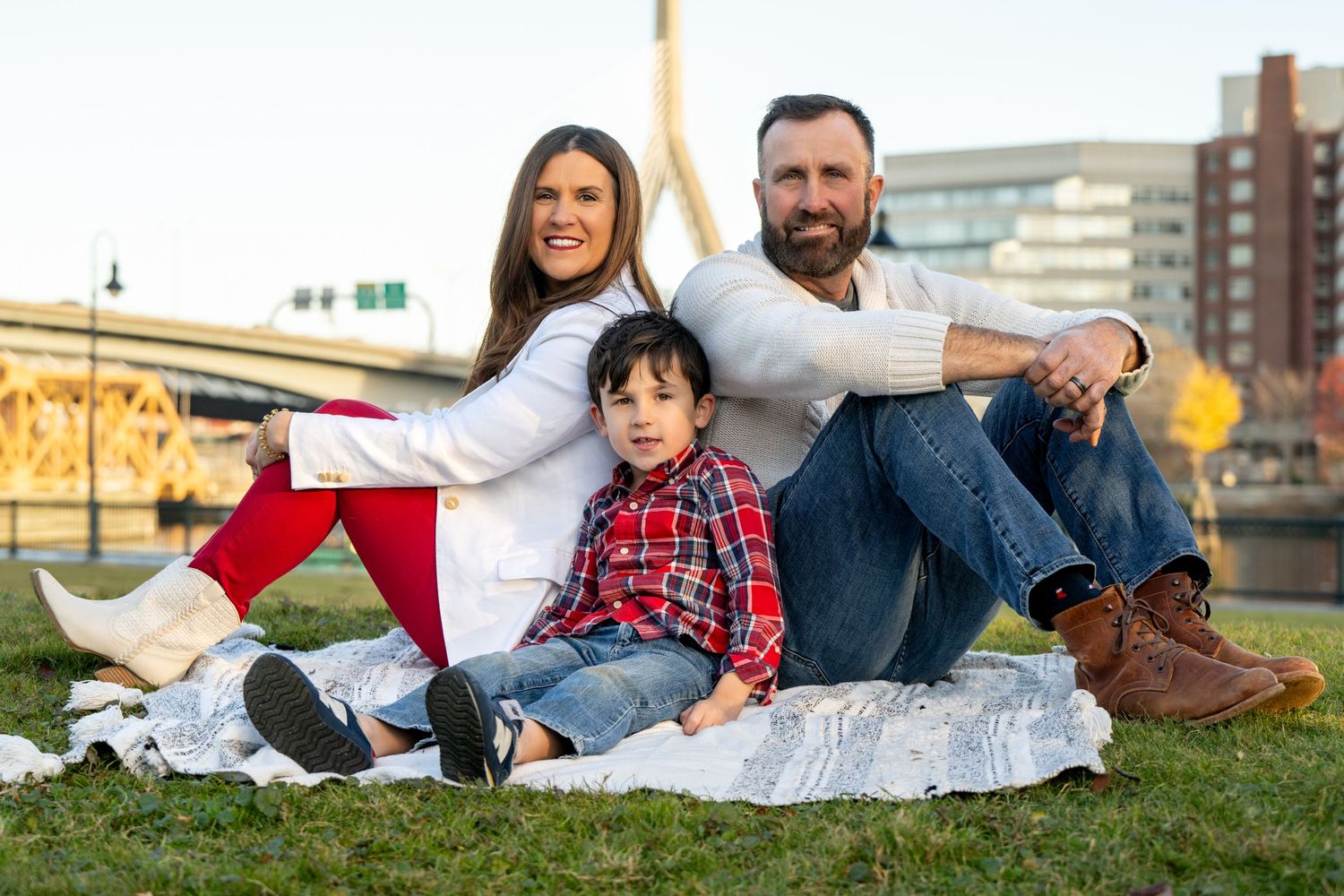 A family sits together on a blanket outdoors on grass near urban buildings during a sunny afternoon.
