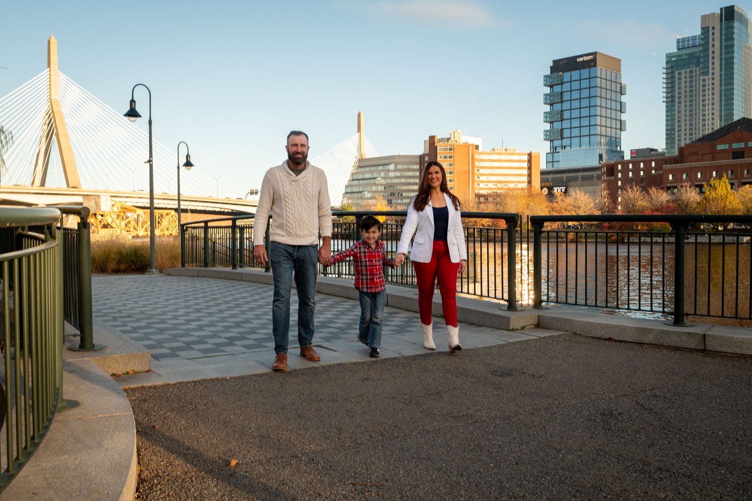 A family walking along Boston's waterfront with the Zakim Bridge and modern buildings visible in the background at sunset.