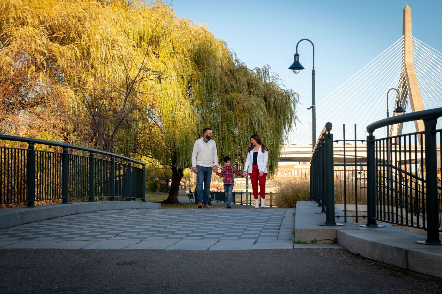 People walking along a waterfront boardwalk with tall bridge and golden willow tree on autumn afternoon.
