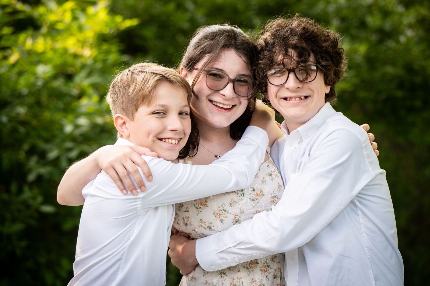 Three people embrace and smile together in a warm outdoor portrait against greenery. Massachusetts Family photographer Serving New England