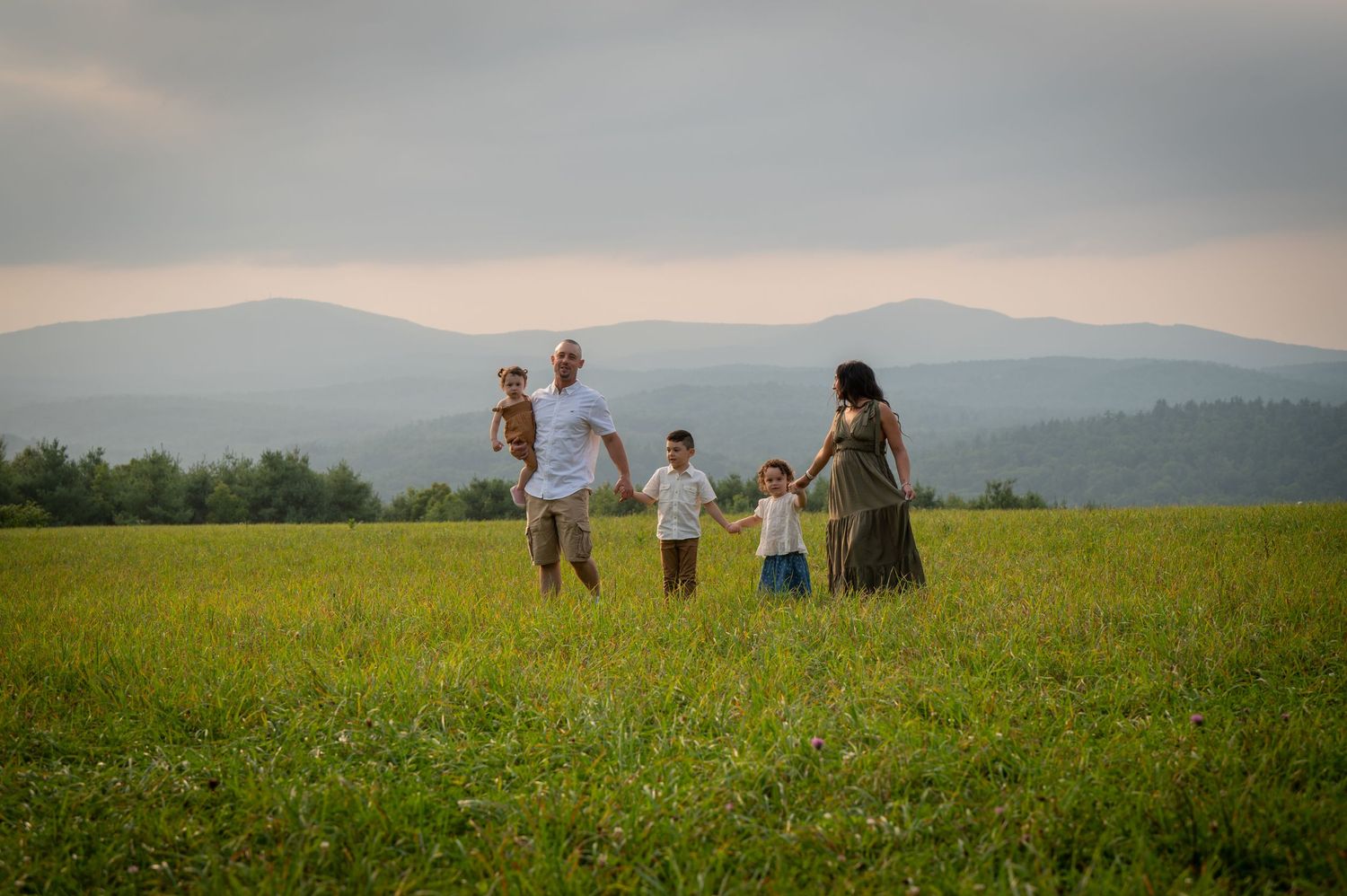 A family holds hands while walking through a sunny mountain meadow at sunset. New Hampshire Family photographer Serving New England